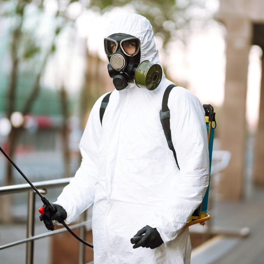 Man in protective suit and mask sprays disinfector onto the railing in the public place. Covid -19.